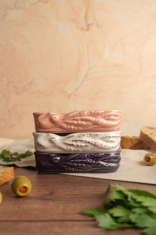 A set of ceramic octopus plates in three different colors: white, lilac, and pink, displayed on a wooden surface with a piece of bread and some olives to the side.