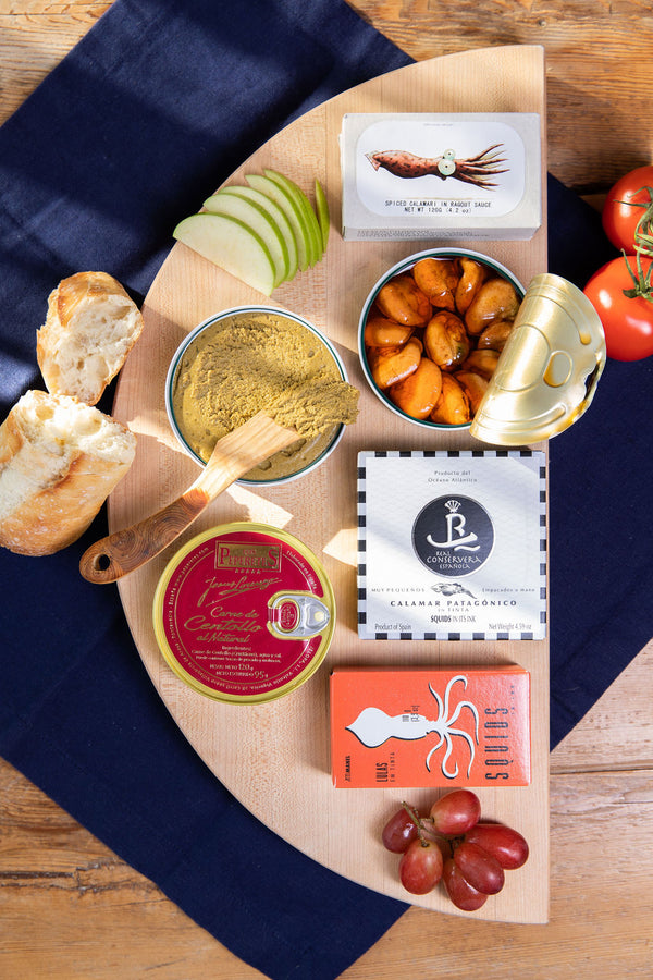 An assortment of gourmet seafood and condiments displayed on a wooden board, including canned lobster, dried seafood, and a small bowl of what appears to be a seafood sauce or spread, accompanied by sliced white bread and grapes.